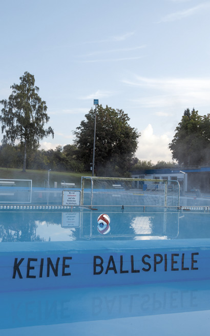 Freibad, umgeben von Bäumen mit Hinweis am Beckenrand ‘Keine Ballspiele’ und Ball schwimmt im Wasser vor einem Wasserballtor